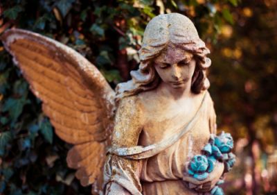 This is a picture of a statue representing an angel. She is holding some blue flowers in her left hand. Is depicted with flowing locks of hair, wings, and her robe has fallen delicately from her right shoulder to rest on her arm.