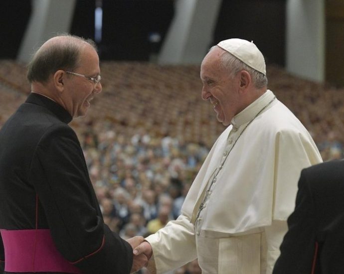 The image shows Pope Francis greeting another religious leader in front of a large crowd in attendance.