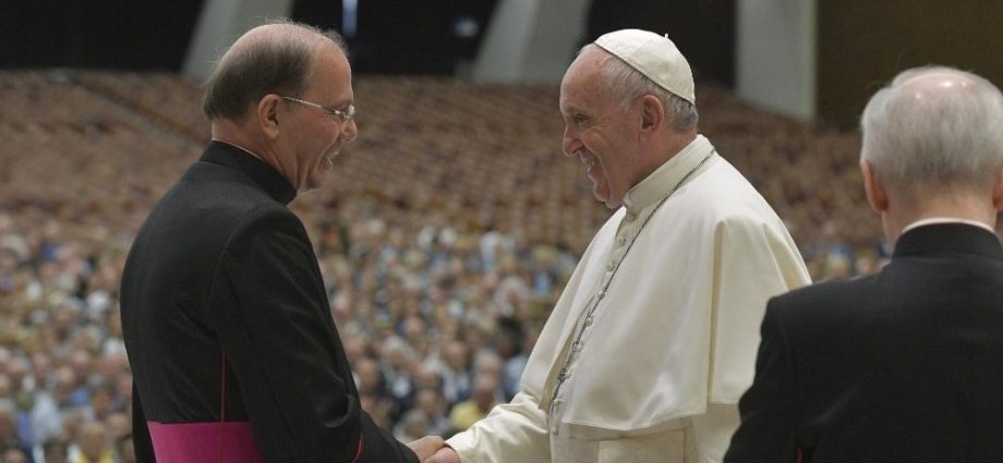 The image shows Pope Francis greeting another religious leader in front of a large crowd in attendance.